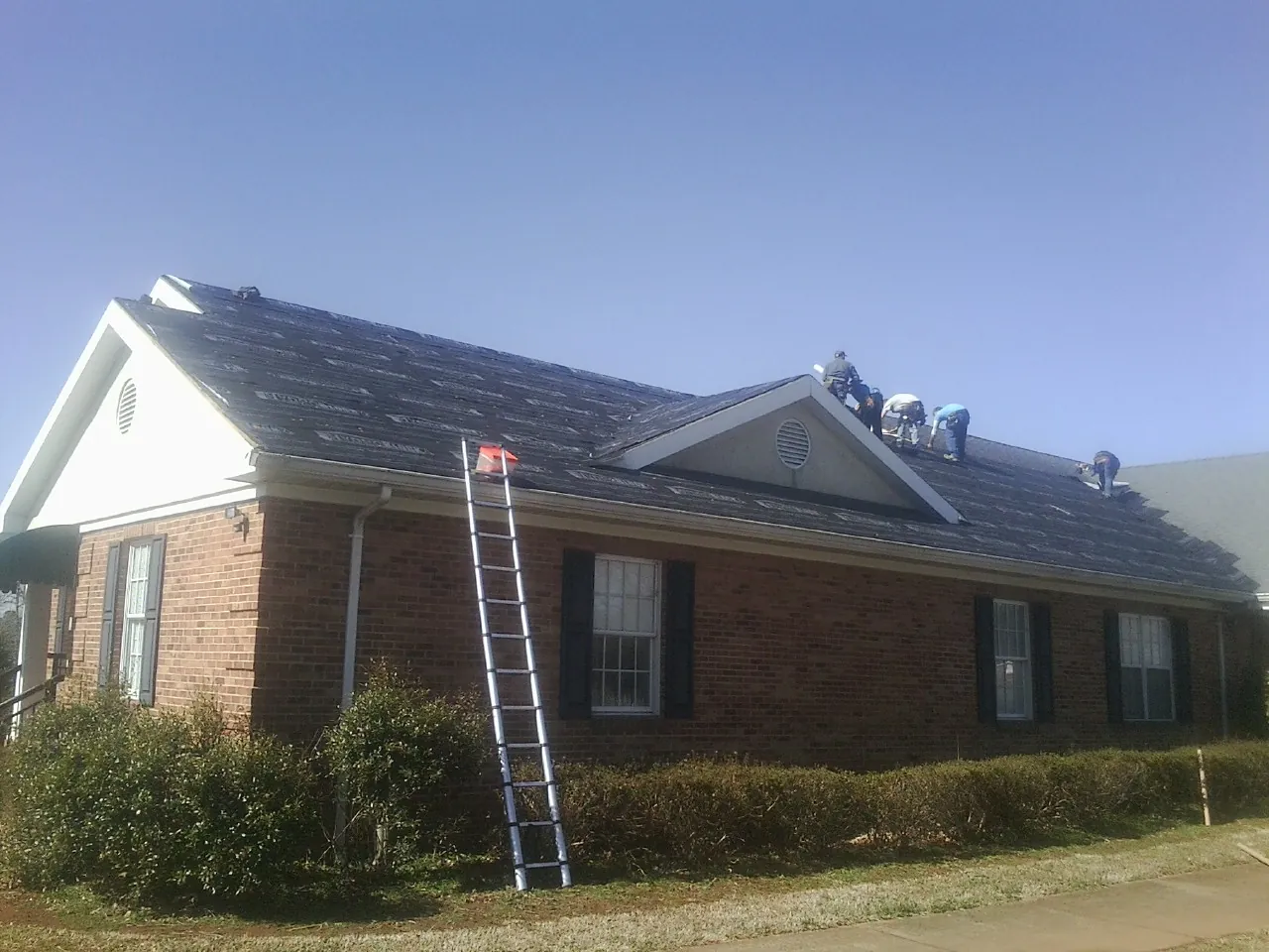 Crew tearing shingles off a brick ranch roof with a ladder set — Emergency Roof Repair work by our crew in Mc Leod