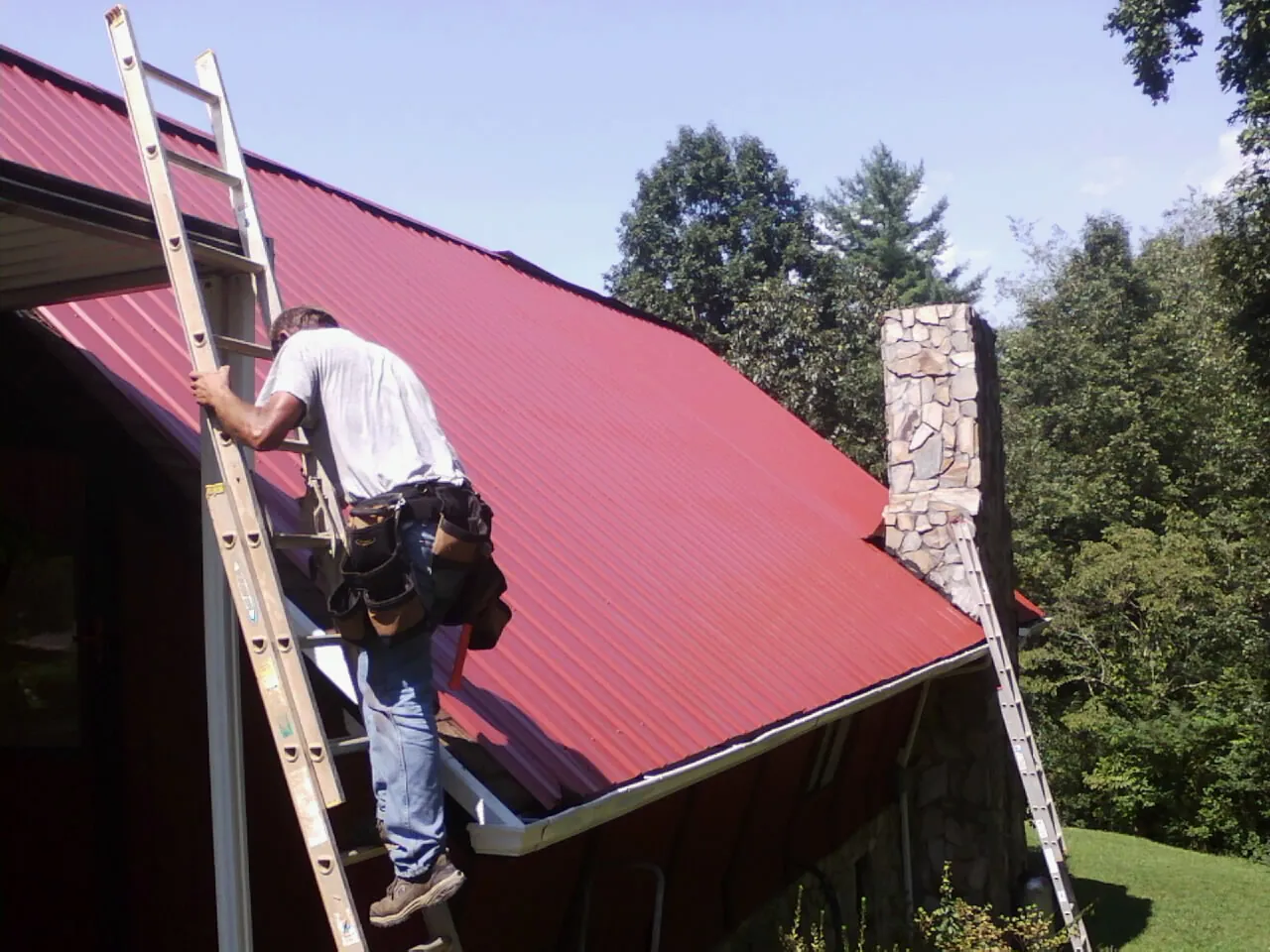 Roofer on a ladder beside a red metal roof and stone chimney — Soffit & Fascia Repair by Roofing Brothers in Mc Leod